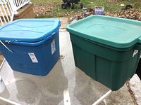 Photo shows two large closed plastic storage totes, one blue and one green, placed on an outdoor glass table.