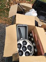 Wide shot showing two cardboard boxes filled with various kitchen ware items including baking pans, egg poacher pan, and wooden cutting board.