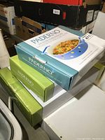 Stacked boxes of three Paderno baking dishes: one blue 9 inch round dish and two green 12 x 8 inch rectangular dishes, all in new condition.