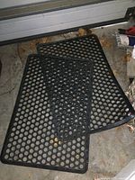 Two black rubber entrance mats with circular hole pattern on dirty concrete floor, showing full products overlapping