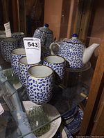 Photo showing the full set of a small teapot with 5 matching cups inside a glass cabinet.