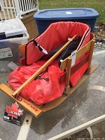 Photo of the wooden child's sleigh with red padded seat, side cushions, and attached red Pelican bag on a glass table with plastic bins in the background.