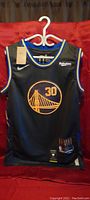 Front view of black Stephen Curry Golden State Warriors jersey with blue and yellow trim, bridge logo and number 30 in gold, hanging on hanger with tags attached.