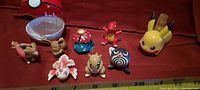 Small Pokemon figures including Eevee, Vaporeon, Sandshrew, Poliwhirl, Charmeleon, and others, plus a yellow Pikachu-themed Pokeball with keyring shown on red fabric with measuring tape in view for size reference.
