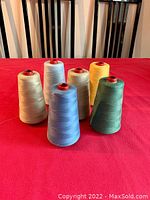 Six spools of cotton thread in various colors including white, blue, yellow, green, and beige arranged in two rows on a red surface with black chairs in background.