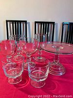 Photo showing 7 glass dessert bowls and the pedestal cake serving platter on a red cloth background with chairs in the background.