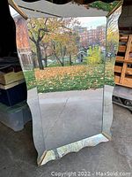 Full view of a large retro floor mirror showing its beveled edges and distinct base shape, standing indoors near an open garage door.