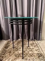 Front view of the side table showing the round frosted blue glass top and black metal base with three legs and circular metal support rings.