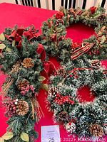 Three Christmas wreaths displayed on a red surface, showing the long wreath with pinecones and red bows, the large circular wreath with red flowers and burlap ribbons, and the medium frosted wreath with pinecones and berries.