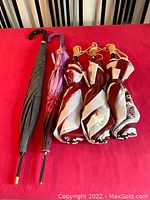 All five umbrellas laid on a red surface showing their handles and closed canopies. Two standard umbrellas with different handles and patterns, three golf umbrellas with maroon and white colors and wooden handles.