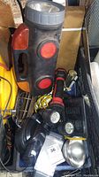Photo showing collection of flashlights of various sizes and styles including a large black flashlight with multiple red reflectors, smaller handheld flashlights, headlamp, and accessories in a container.