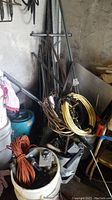 Stack of metal rods, angle irons, and metal sheets against wall with coils of wiring and cables in front, piled on buckets and floor.