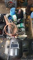 Wide view showing welding helmets on bottom and crate containing welding rods, wires, and other accessories piled on top.