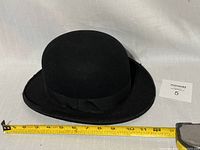 Front view of black bowler hat on white surface with measuring tape showing about 11.5 inches diameter
