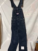 Full view of dark wash denim overalls with adjustable straps hanging against a white backdrop.