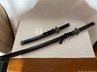 Lot of two black ninja swords laid flat on a white surface showing full length and sheaths.