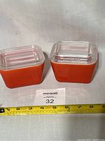 Two square light red Pyrex refrigerator dishes with clear glass lids placed side by side with a measuring tape in front showing around 4 to 5 inches width