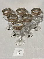 Set of six clear wine glasses with wide 24k gold encrusted rims arranged in two rows on white cloth.