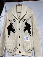 Front view of the cream wool knit sweater with two horse head motifs, tortoise buttons, shawl collar and front pockets showing knit details.