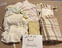 Photo shows six folded bed sheets in different colors and patterns arranged on the floor with a sign reading 'LOT #48 Bed Sheets'.