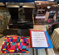 Photo showing all items in the lot: two metal Bluetooth speakers, LG 5-disc DVD player with remote, Keurig K-Supreme in box, two computer arm rests, two binders, and a colorful cloth