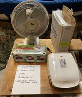 Photo showing four items on a wooden table: a white standing table fan, a boxed booster fan, a boxed grilling machine, and a white wall panel heater. Handwritten note describes lot contents.