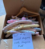 Photo showing a box containing multiple wooden and cloth-covered clothing hangers in various colors and patterns, with a paper note indicating the lot is wood and cloth hangers.