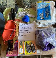 Overview of lot showing white heating blanket with remote control, red boot dryer, deflated purple exercise ball, various hair brushes including round and paddle styles, vacuum cleaner bags and cleaning brushes, and handwritten note describing items.