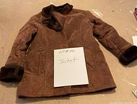Photo of a brown shearling jacket laid flat on the floor showing collar, cuffs, button closure, and pockets with a card labeled 'LOT# 116 Jacket'.