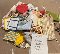 Photo shows a pile of mixed kitchen textiles including multiple gloves, towels, oven mitts, and pot holders in several colors and patterns. A handwritten paper lists Lot #109 kitchen supplies, gloves, towels.