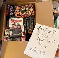 Photo of an open box filled with DVD and CD movie cases, includes visible titles and a handwritten sign indicating 'Lot #67 DVD/CD Full Box Movies'.