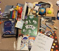 Top-down photo of various office and school supplies arranged on floor including clipboards, globe, stationery, decks of cards, pens, pencils, stapler, erasers, and small tennis ball.