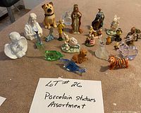 Photo of 18 assorted porcelain and glass statues and figurines arranged on brown surface including animals and human busts.