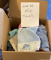 Box of assorted mixed towels including light blue towel with lace and flower embroidery detail, blue, green, coral, and beige towels.
