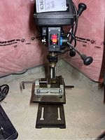 Full view of Mastercraft bench drill press showing black top housing with laser and drill handle, metal vertical support, adjustable worktable, and base with mounted metal vise.