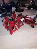 Angled view showing three red axle stands and collapsed MotoMaster hydraulic jack on concrete floor