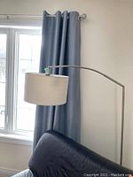 Full view of the floor lamp showing its metal arched arm and beige fabric shade in front of a window with curtains and next to a black chair.