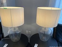 Two table lamps with hammered glass bases and white fabric drum shades placed on a black table.