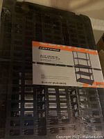 Top view showing shrink-wrapped black shelving unit in original packaging with manufacturer label and dimensions.