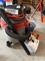 RIDGID wet/dry vac showing vacuum hose, orange extension cord wrapped on top, gray bucket, and red motor housing with RIDGID branding.