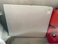 Photo of folded white plastic table resting upright against a wall showing the surface and sizing.