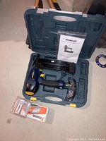 Full kit open in case showing Mastercraft finishing nailer in blue and black with rubber grip, plastic wrapped accessory or goggles, instruction manual, and packs of nails.