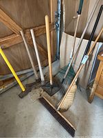 Full view of all tools standing and leaning against a gym-style door, including push broom, corn broom, rakes, pickaxes, tamper, and sledgehammer