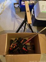 Wooden plank mounted on black metal tripod base with box of orange and black bar clamps underneath