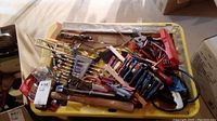 Top view of mixed hand tools including wrenches, screwdrivers, wire cutters, hammer head, chisels, and drill bits in a yellow tray