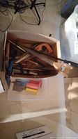 Photo showing a box containing multiple handsaws with wooden and plastic handles, a crowbar, and other hand tools not individually detailed.