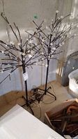 Two black metal decorative lighted trees with white flower lights on their branches standing against a cinder block wall. The wiring is coiled at the base on the floor.