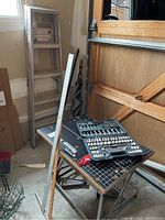 Workbench with black surface and orange edges, socket set case opened on top, wooden yardstick leaning against it, five metal step ladders arranged behind workbench
