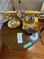 Two vintage rotary dial telephones on a wooden round table, one cream-colored and one yellow.