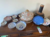 Full view of assorted Oriental ceramic dishes and decor on a wooden surface, showing tureen, bowls, small plates, a black decorative vase, and a blue floral plate.